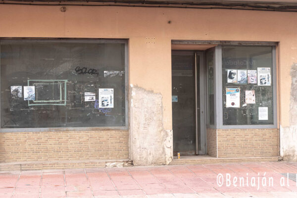 Fachada de un comercio cerrado en Beniaján, con cristales sucios y carteles de publicidad pegados, mostrando un local abandonado y sin actividad.