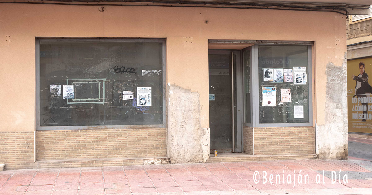 Fachada de un comercio cerrado en Beniaján, con cristales sucios y carteles de publicidad pegados, mostrando un local abandonado y sin actividad.