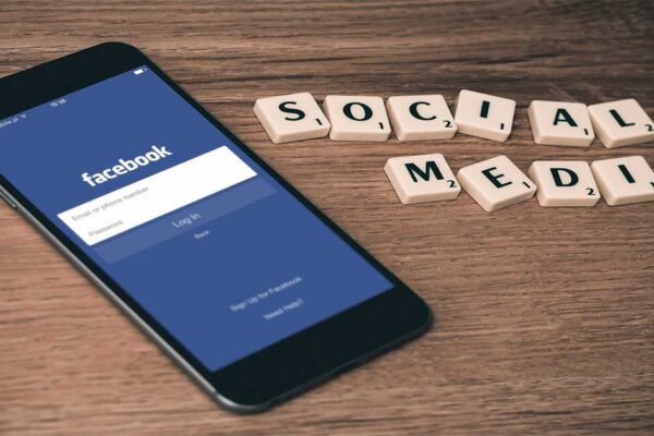 Teléfono móvil mostrando la pantalla de inicio de Facebook sobre una mesa con fichas de dominó que forman la frase “Social Media