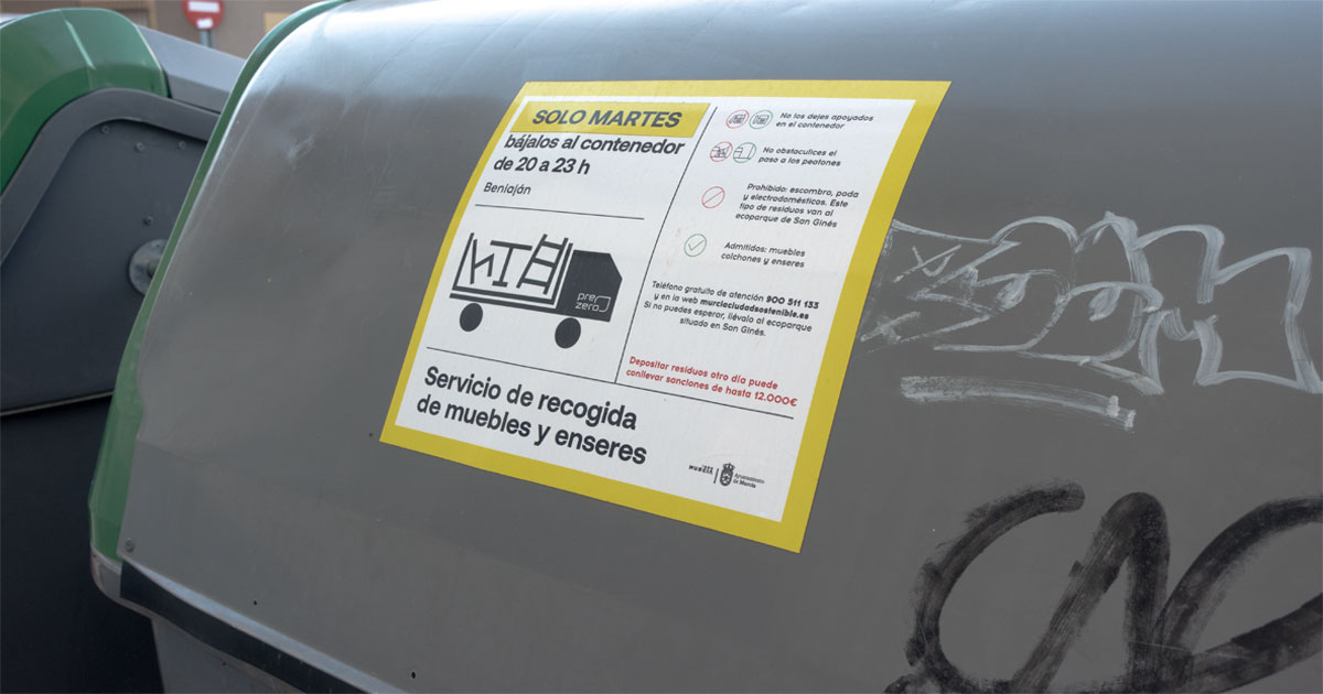 Cartel de aviso con el estricto horario de recogida de muebles y enseres frente a un grupo de contenedores en Beniaján.