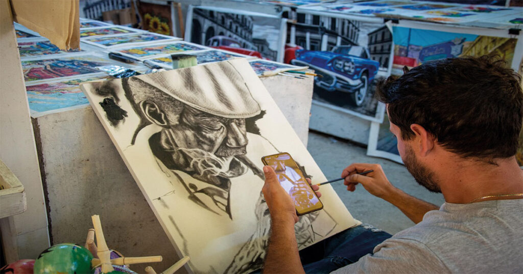 Persona pintando un cuadro inspirándose en una foto en su móvil, rodeada de láminas y materiales de pintura en un espacio creativo