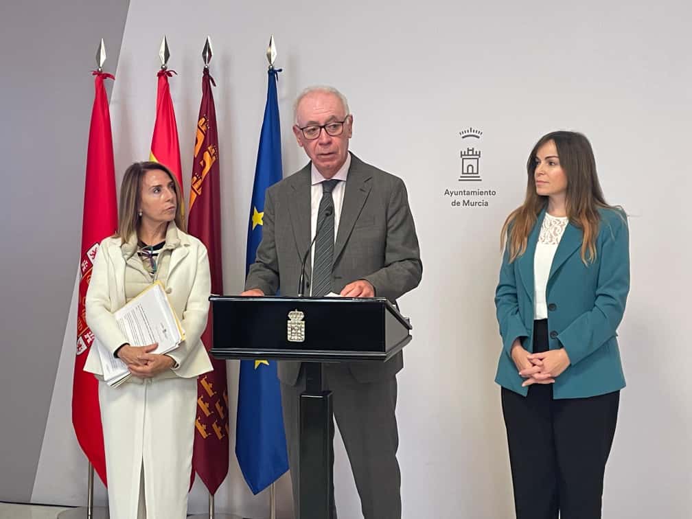 Antonio Navarro JG, en el atril del Ayuntamiento de Murcia con el escudo del Consistorio, acompañado por dos políticas durante la presentación de la medida de compensación por la DANA