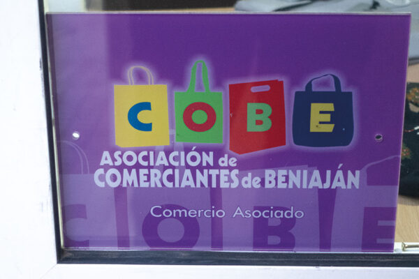 Detalle de la pegatina morada de la Asociación de Comerciantes de Beniaján (COBE) con cuatro bolsas de compra representando las letras del acrónimo, en la fachada de un comercio local.
