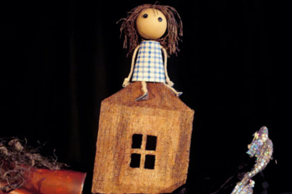 Muñecos y títeres de La Canica Teatro en el Auditorio de Beniaján, con una muñeca sentada en una casita de madera y objetos de utilería sobre fondo negro