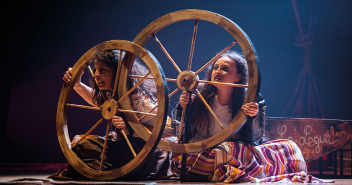 Dos actrices de Yampo Teatro vestidas de época sujetan simbólicamente ruedas de carro antiguas en el escenario, representando el viaje a través de la historia femenina del teatro español en la obra "¡Mujer tenías que ser!" en Beniaján.