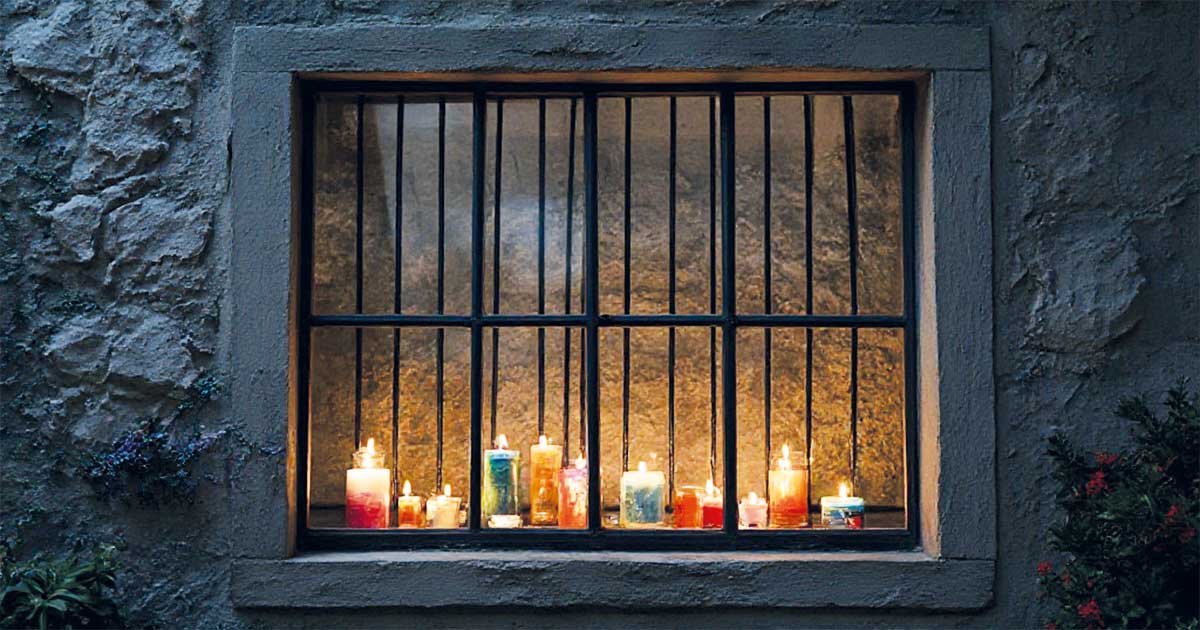 Fachada de edificio antiguo en Beniaján iluminada por múltiples velas de colores durante el evento cultural Beniaján en Vela 2025, con ventana y rejas visibles.