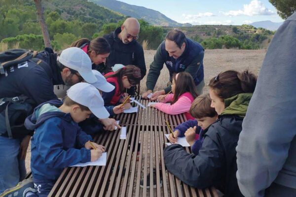 Grupo de niños sentados en una mesa en el parque Majal Blanco realizando actividades educativas de la Escuela de Naturaleza de Navidad, acompañados por monitores del Ayuntamiento de Murcia