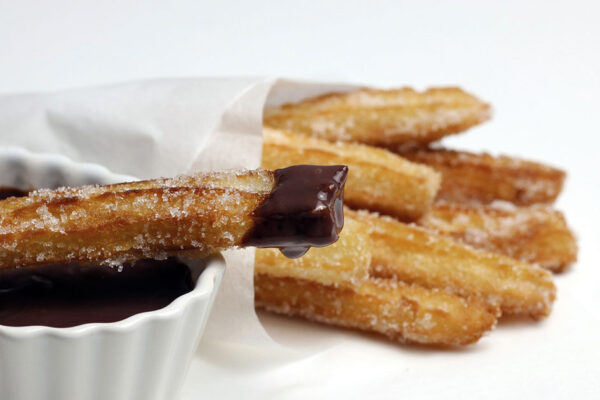 Churros en su papel sobre fondo blanco, con un churro colocado sobre una taza con chocolate caliente y la punta del churro mojada en chocolate