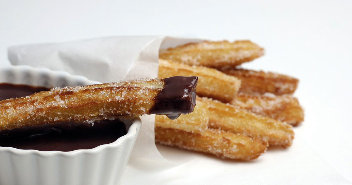 Churros en su papel sobre fondo blanco, con un churro colocado sobre una taza con chocolate caliente y la punta del churro mojada en chocolate