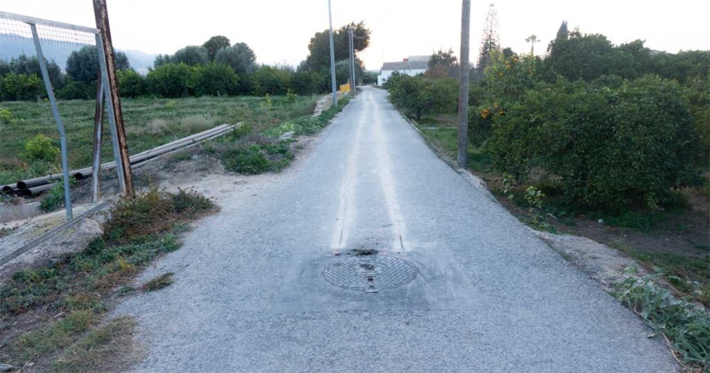 Tramo del Carril Giles en Beniaján con marcas de corte para las obras de bombeo; se observan tuberías de PVC negro al lado izquierdo junto a un registro en la vía.