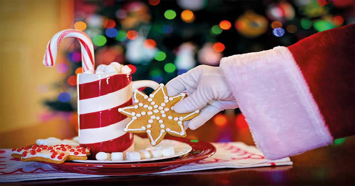 Mano de Santa Claus colocando un adorno navideño sobre una mesa con centros decorativos, ilustrando la Navidad en barrios y pedanías del Ayuntamiento de Murcia.