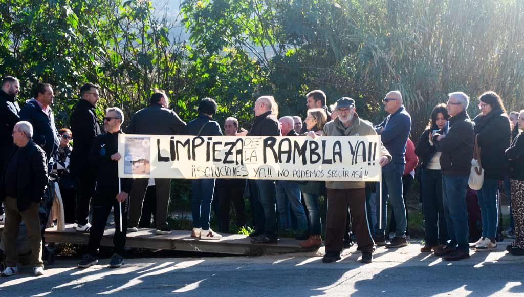 Grupo de asistentes con pancarta y lema en la concentración por la limpieza de la rambla del Garruchal en El Secano, con políticos presentes al fondo.