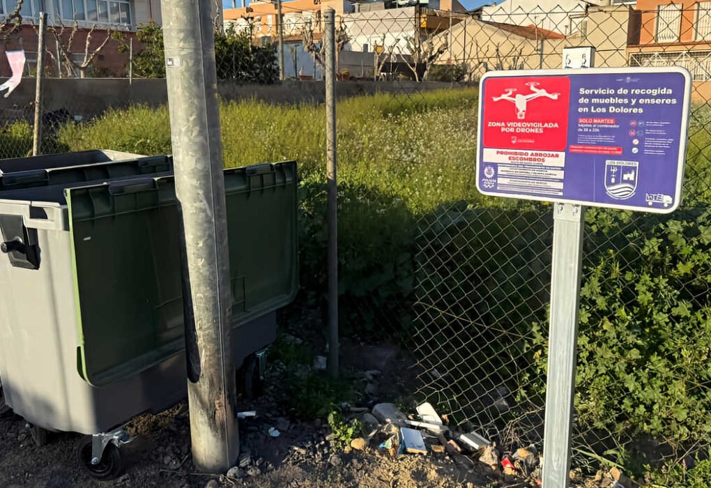 Contenedor de residuos junto a un solar en Los Dolores con un cartel de videovigilancia mediante drones de la Junta Municipal.