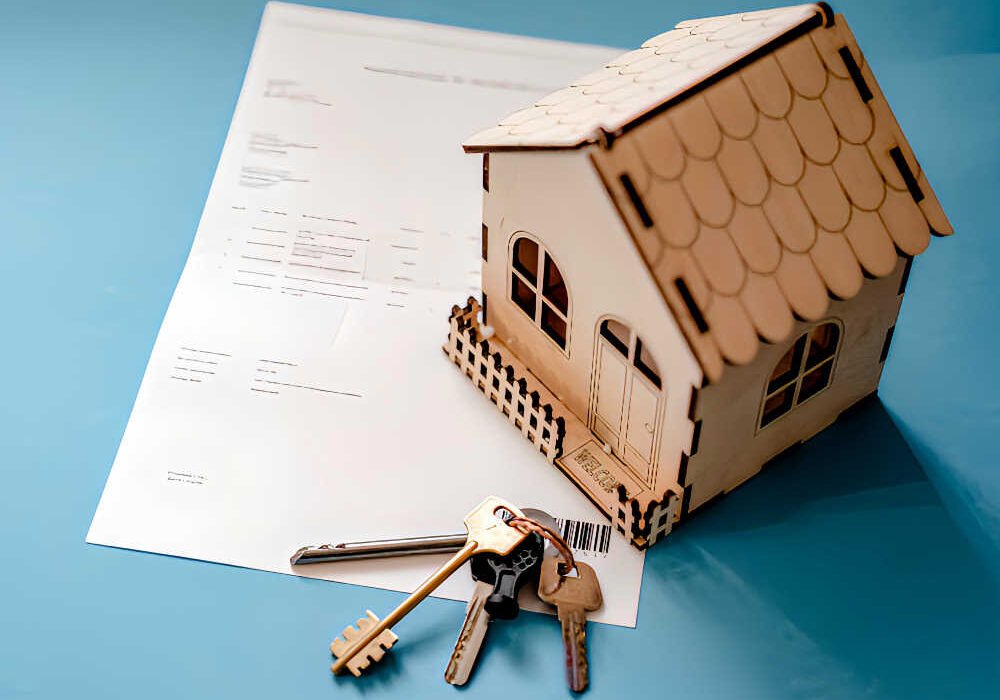 Maqueta de casa de madera, llaves y documento de contrato de alquiler de vivienda sobre fondo azul.
