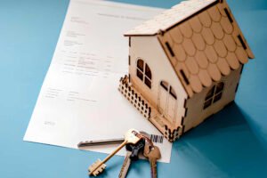 Maqueta de casa de madera, llaves y documento de contrato de alquiler de vivienda sobre fondo azul.
