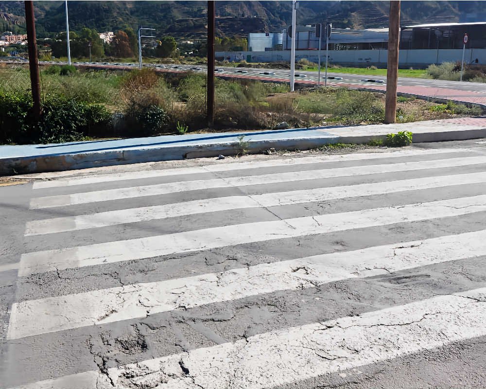 Paso de peatones en calzada deteriorada que desemboca contra un bordillo elevado azul y postes de madera en San José de la Vega.
